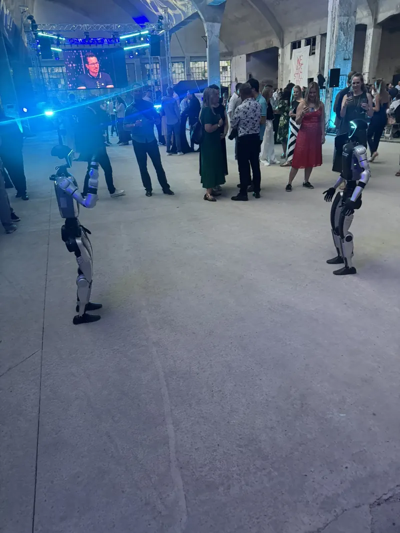 Dva humanoidi mezi hosty v osvětlené hale